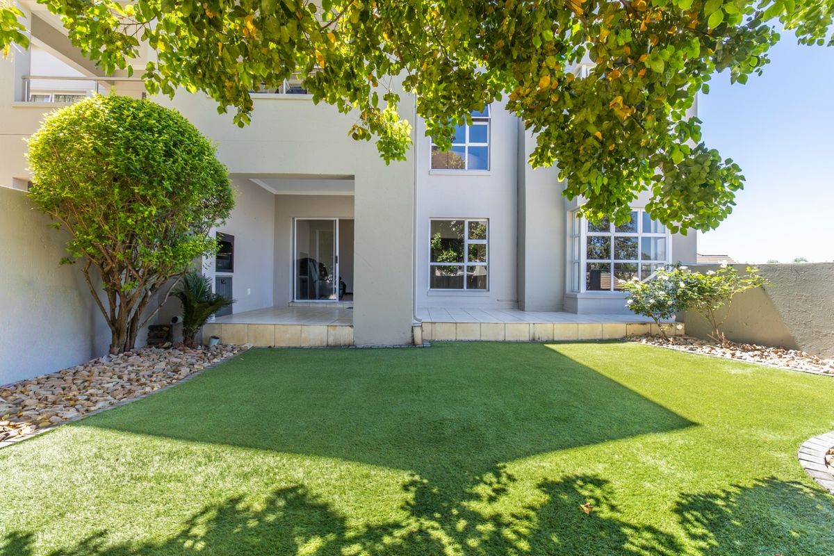 Rear view of the apartment featuring a covered patio with a built-in braai, sliding doors, and a well-maintained artificial lawn garden.