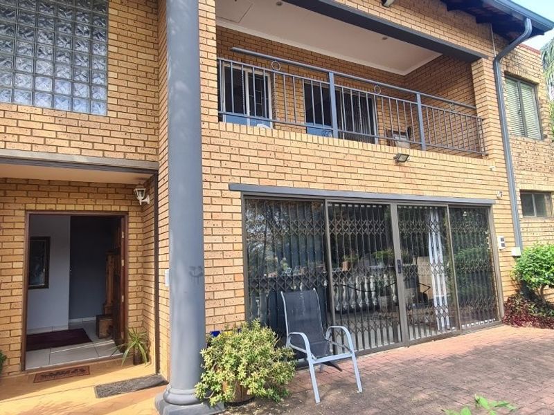 Rear exterior view of a double-story face brick house featuring a balcony with metal railings, sliding glass doors, and a paved patio area.