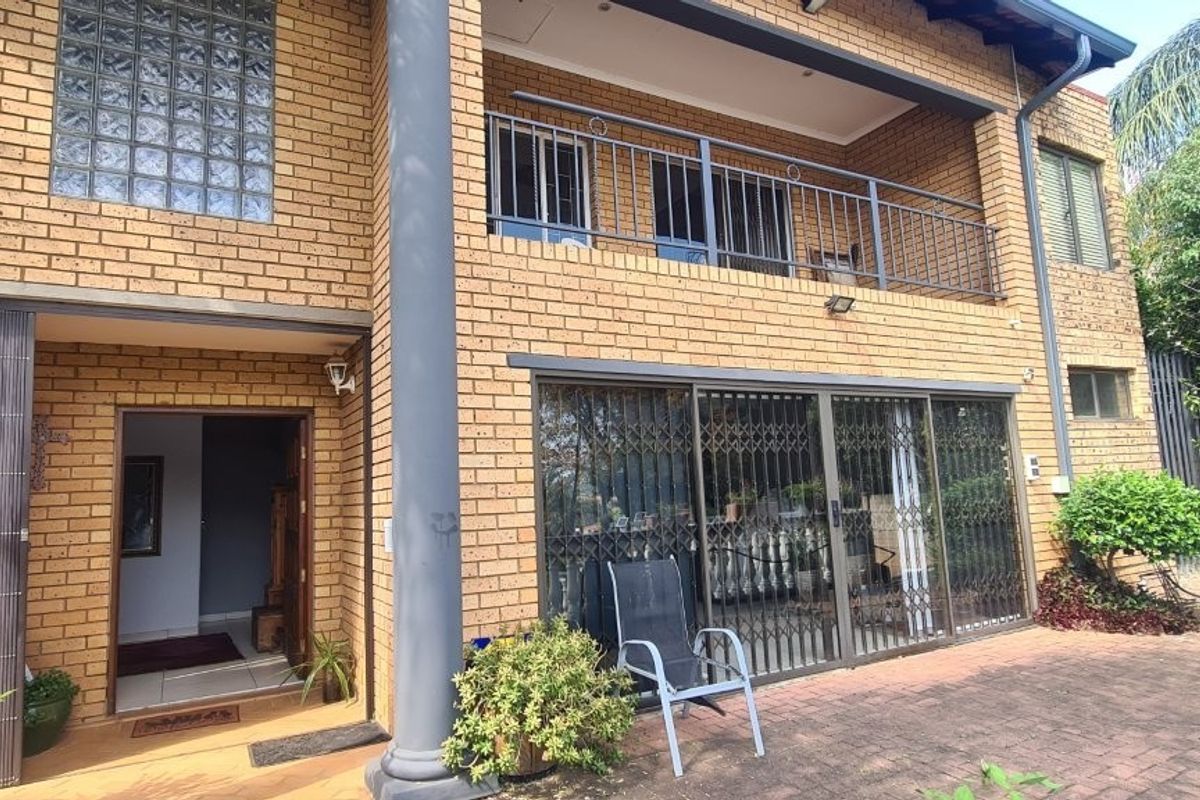 Rear exterior view of a double-story face brick house featuring a balcony with metal railings, sliding glass doors, and a paved patio area.