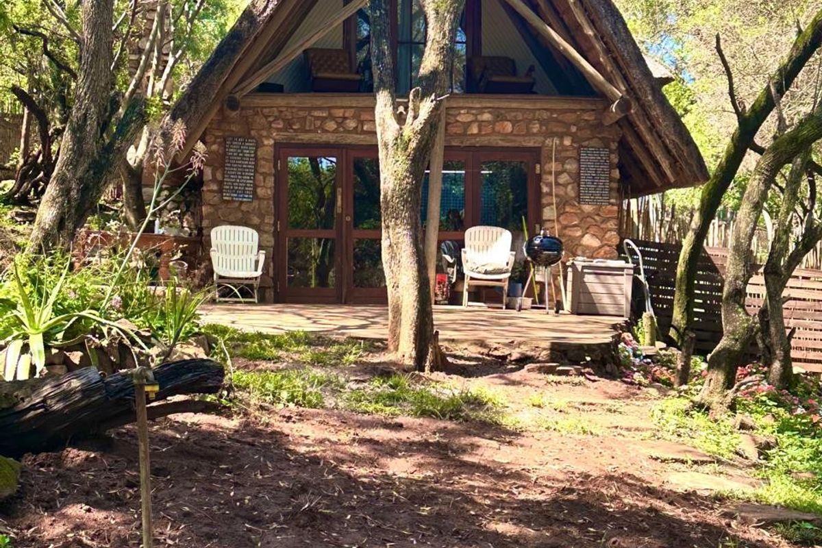 A charming A-frame house featuring stone cladding, a thatched roof, and a wooden deck nestled in a wooded garden setting.