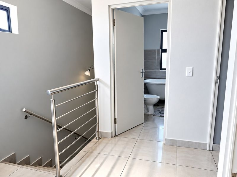 A bright, modern hallway featuring tiled flooring and a sleek stainless steel balustrade, providing access to an adjacent bathroom.