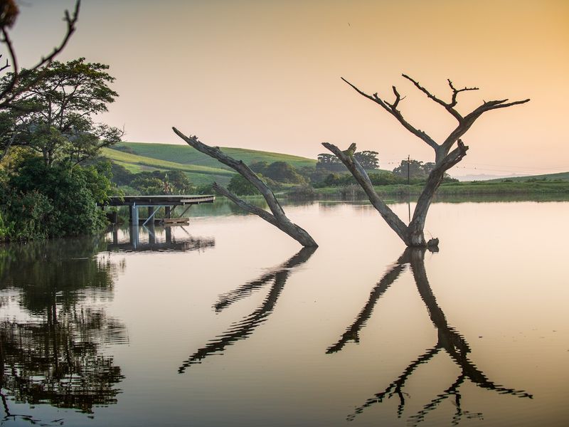 A scenic view of a tranquil dam on the small holding, featuring a wooden jetty and natural landscape surroundings at sunset.