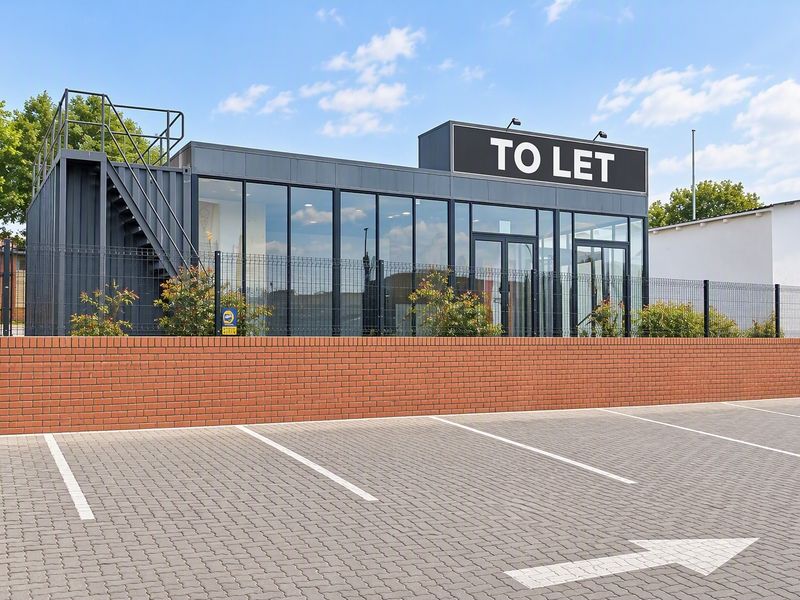 Modern commercial container-style building featuring a full-height glass facade, external staircase, and dedicated paved parking area with brick boundary wall.
