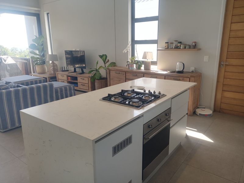 Modern open-plan living area featuring a central kitchen island with a built-in gas hob and oven, complemented by tiled flooring and natural light.
