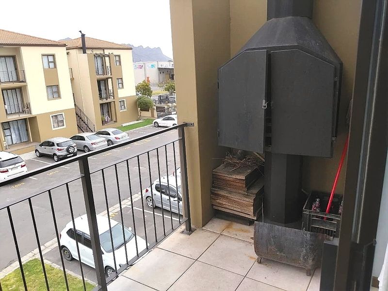 A balcony featuring a built-in braai, tiled flooring, and a metal balustrade, overlooking the apartment complex parking area.