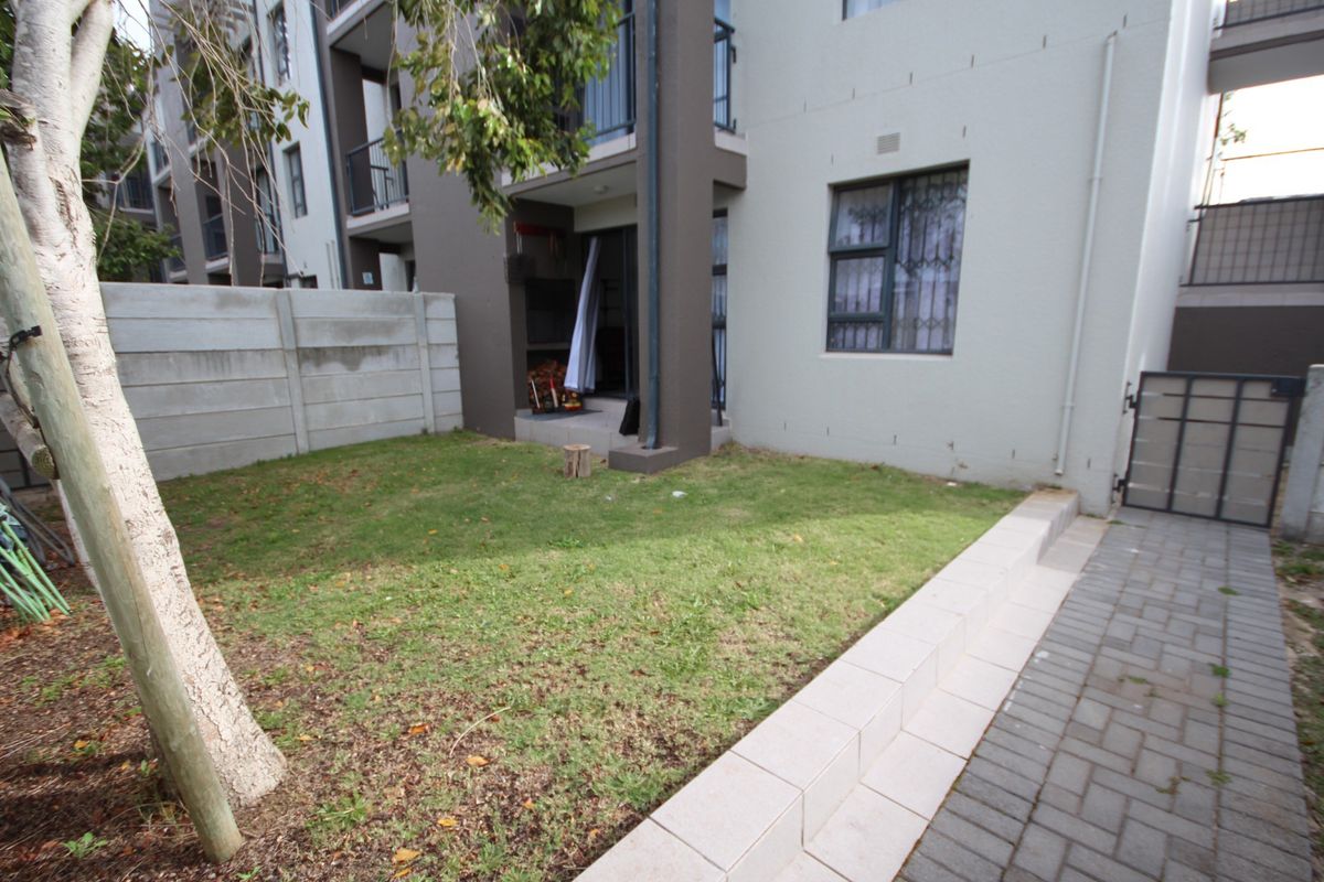A private rear garden area featuring a small lawn, paved walkway, tiled retaining wall, and direct access to the apartment via a sliding door.