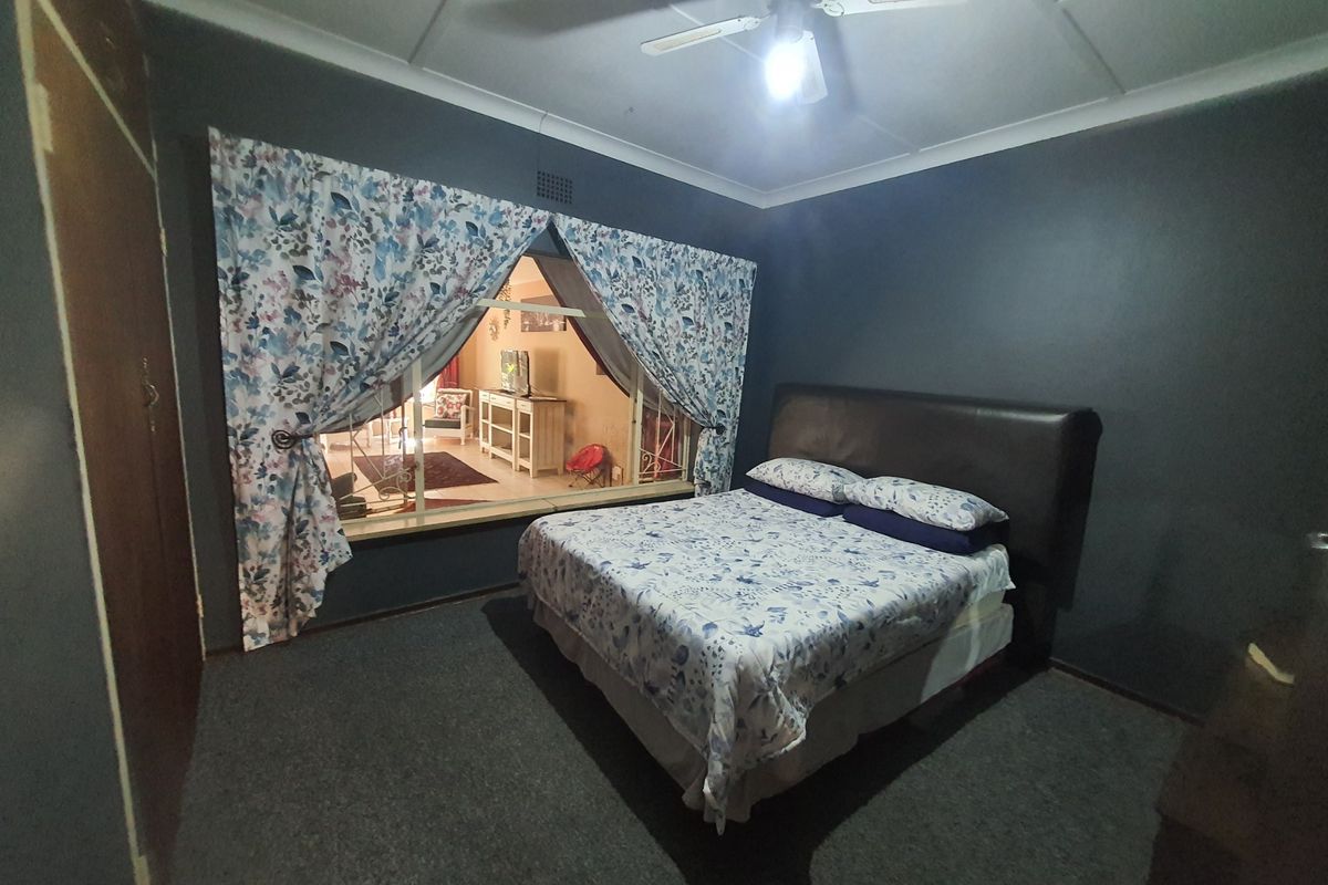 A bedroom featuring dark painted walls, a built-in cupboard, a ceiling fan, and a large window looking into an adjacent living space.