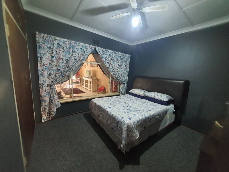 A bedroom featuring dark painted walls, a built-in cupboard, a ceiling fan, and a large window looking into an adjacent living space.