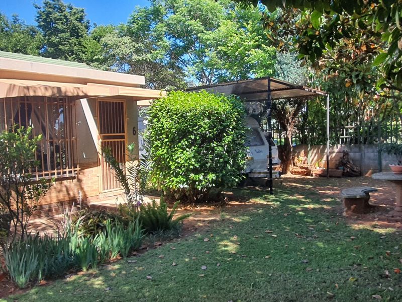 Rear garden area featuring a covered carport, established landscaping, and a permanent stone outdoor seating area adjacent to the house.