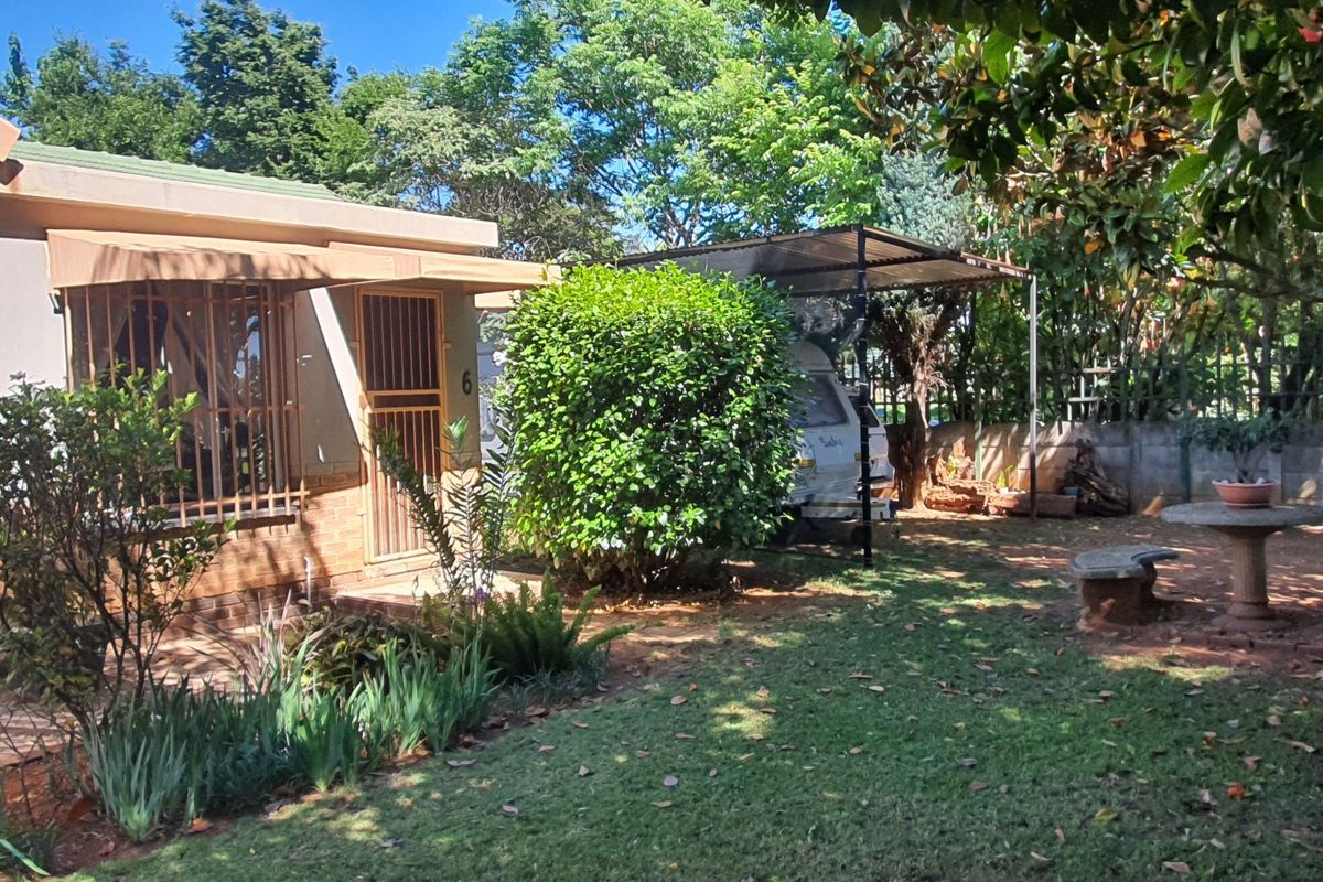 Rear garden area featuring a covered carport, established landscaping, and a permanent stone outdoor seating area adjacent to the house.