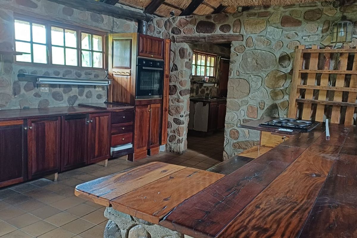 Rustic farmhouse kitchen featuring stone walls, exposed timber beams, thatch ceiling, and traditional wooden cabinetry with integrated oven and hob.