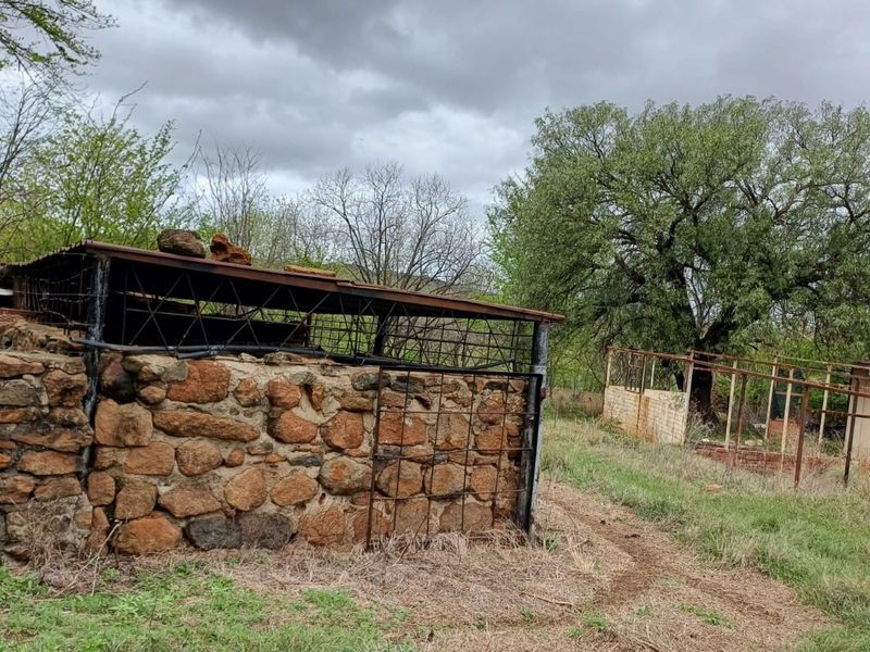 Exterior view of a rustic farm outbuilding featuring stone masonry walls and a weathered metal roof structure, set within a rural landscape.