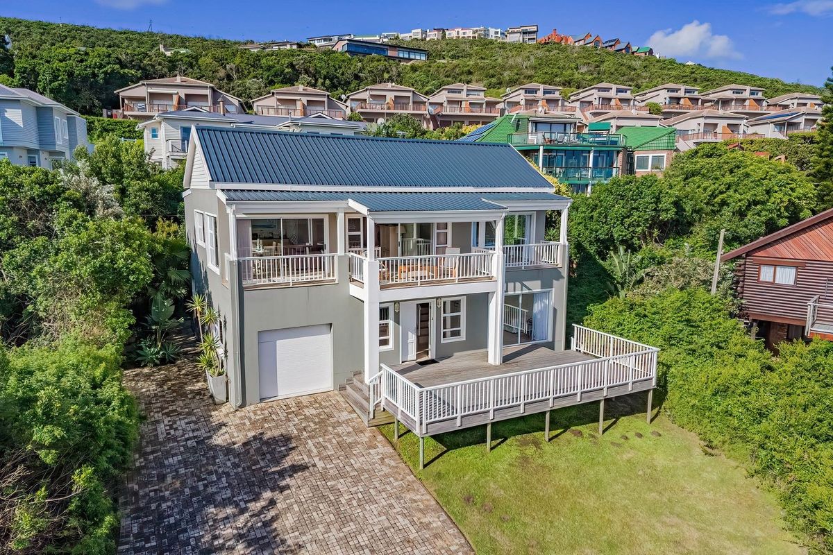 Rear view of a two-story house featuring a metal roof, a spacious timber deck, a balcony, and a paved driveway set within a landscaped garden.