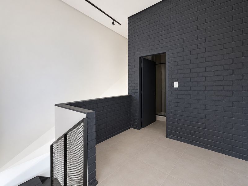 A modern, minimalist hallway featuring a striking dark exposed brick wall, sleek metal mesh balustrade, and neutral tiled flooring.