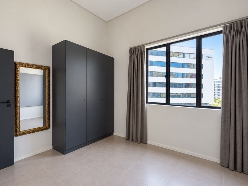 A bright, modern bedroom featuring neutral tiled flooring, a sleek dark built-in wardrobe, and large windows with curtains.