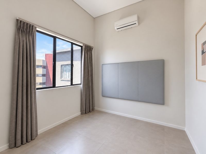 A bright, empty bedroom featuring neutral tiled flooring, a split-system air conditioner, and a large window with installed curtain tracks.