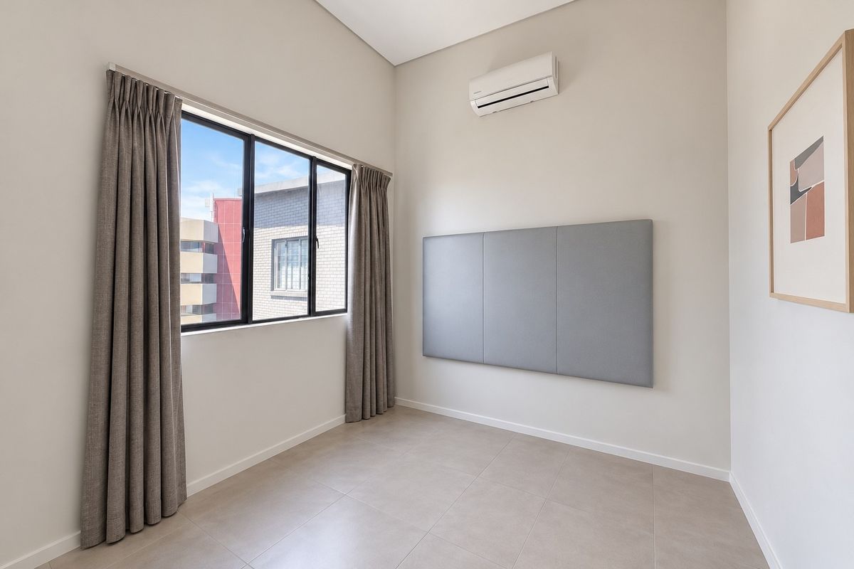 A bright, empty bedroom featuring neutral tiled flooring, a split-system air conditioner, and a large window with installed curtain tracks.
