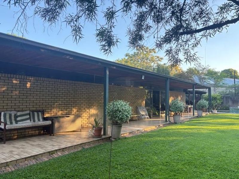 A spacious covered patio featuring brick walls, tiled flooring, and a timber ceiling, overlooking a well-maintained garden area.