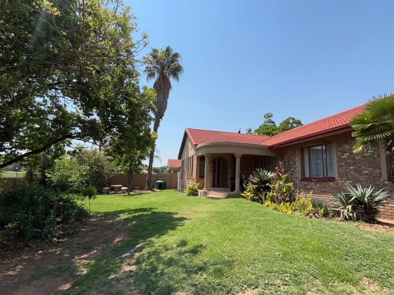 A rear view of a brick house featuring a tiled roof, a covered patio area, and a well-established garden with mature trees.