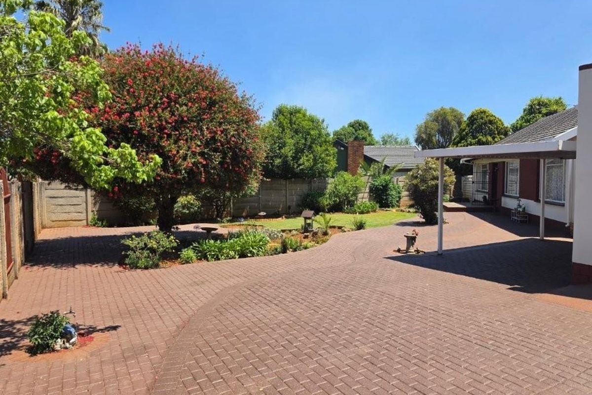 A spacious rear exterior area featuring extensive brick paving, a covered patio space, and a well-maintained garden with mature trees and boundary walling.