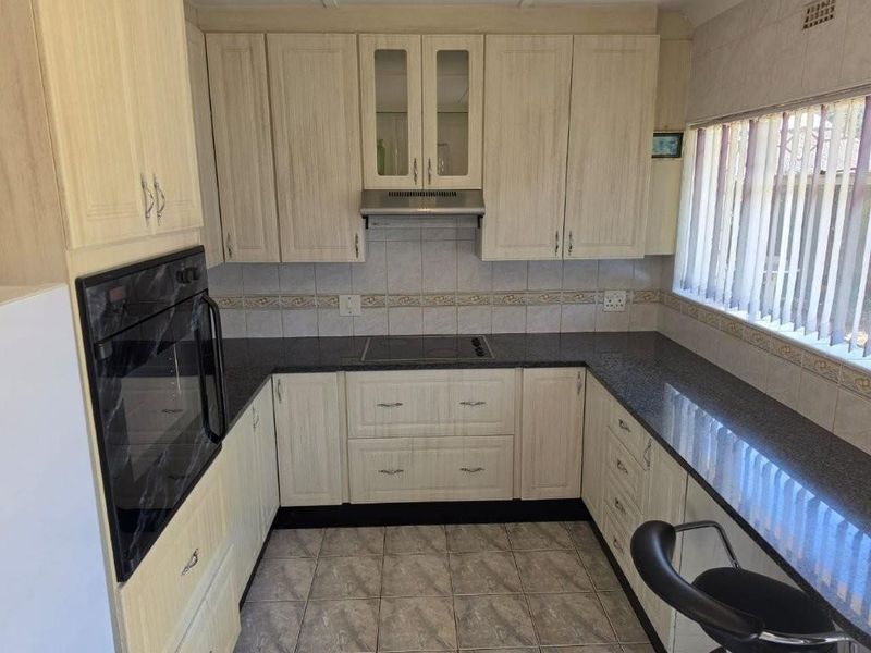 A functional kitchen featuring light wood-grain cabinetry, dark granite countertops, a built-in oven, electric hob, and tiled flooring with a window for natural light.