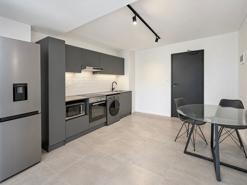 Modern open-plan kitchen featuring sleek dark grey cabinetry, integrated appliances, subway tile backsplash, and contemporary track lighting, seamlessly connected to the dining area.