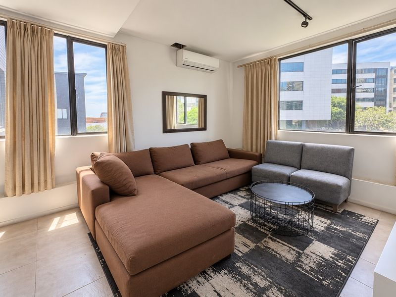 A bright and spacious living room featuring tiled flooring, large windows providing ample natural light, and a wall-mounted split-system air conditioner.