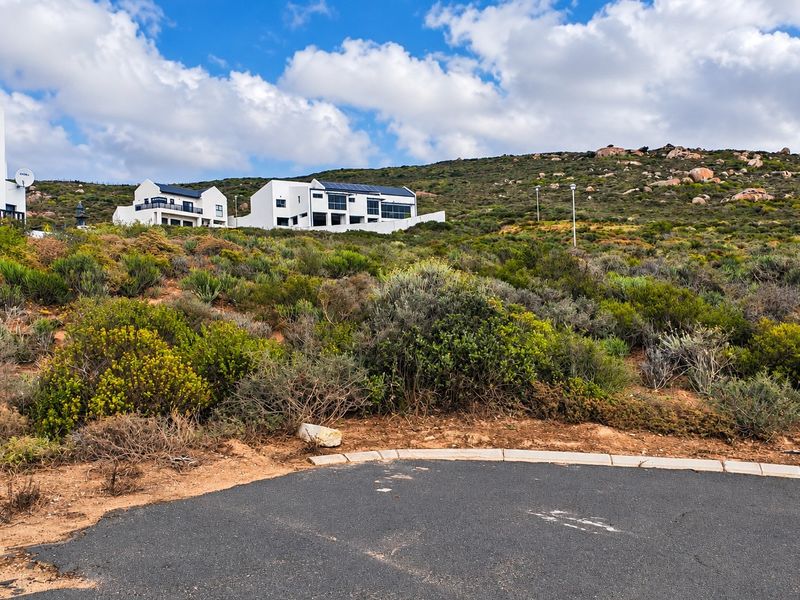 A view of a vacant residential plot featuring natural coastal vegetation, paved road access, and kerbing, with established homes visible in the background.