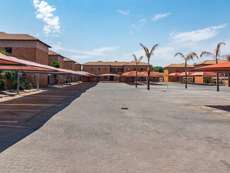 A spacious paved parking area featuring multiple covered carport structures and brick residential buildings under a clear sky.