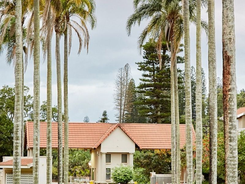 A well-maintained gated entrance to the apartment complex, featuring a security gatehouse, paved driveway, and mature palm trees.
