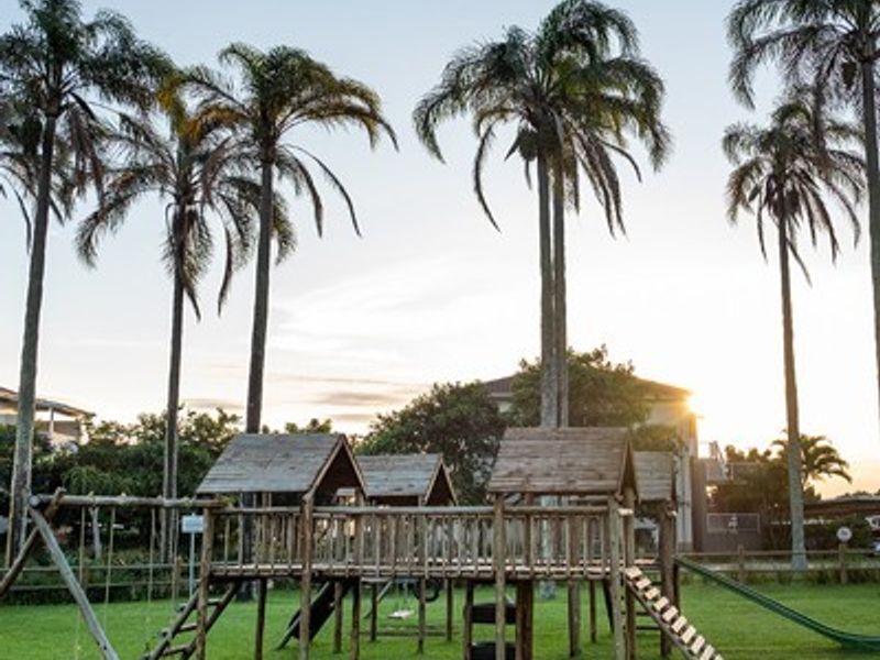 A well-maintained communal garden area featuring a wooden playground structure, swings, and a slide set amongst mature palm trees.
