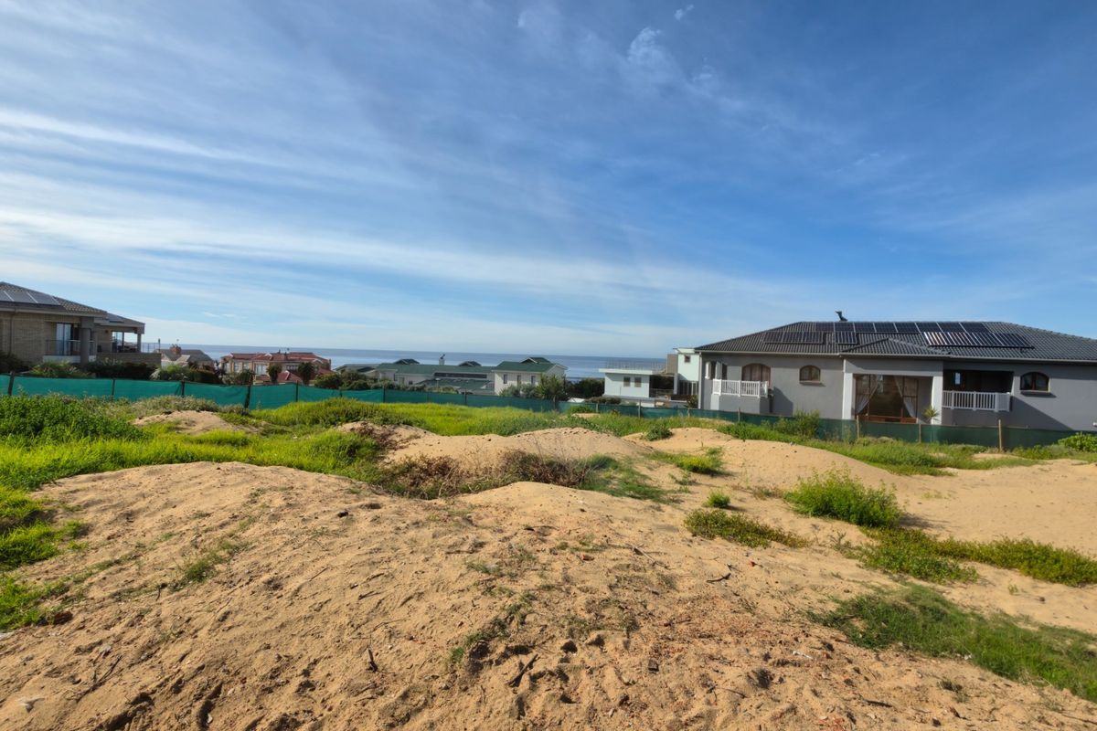 A vacant residential plot featuring sandy terrain and green perimeter fencing, offering a clear view of the surrounding coastal neighborhood.