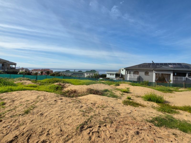 A vacant residential plot featuring sandy terrain and green perimeter fencing, offering a clear view of the surrounding coastal neighborhood.