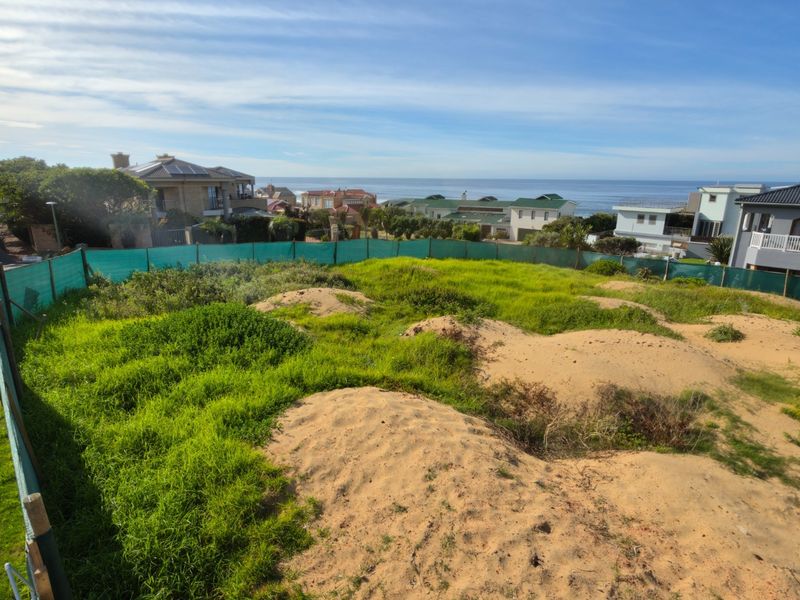 A vacant residential plot featuring natural sandy terrain and coastal views, enclosed by perimeter fencing in a developed neighborhood.