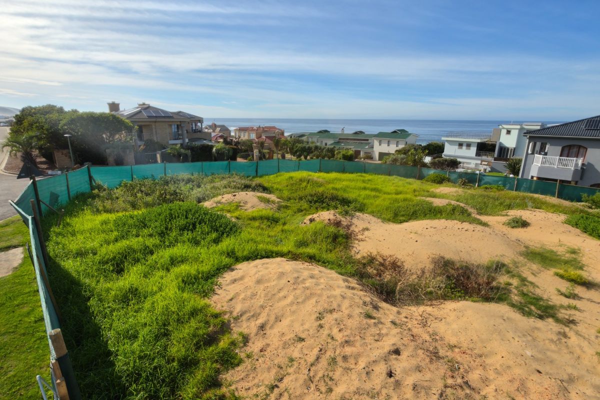 A vacant residential plot featuring natural sandy terrain and coastal views, enclosed by perimeter fencing in a developed neighborhood.