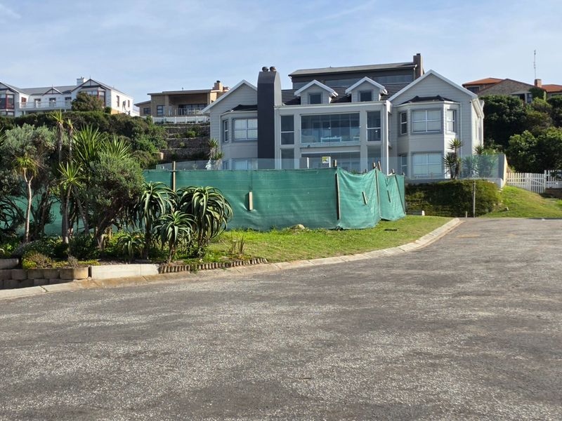 A view of a residential property from the street, featuring a paved driveway, landscaped garden, and a modern multi-story house with a glass balustrade.