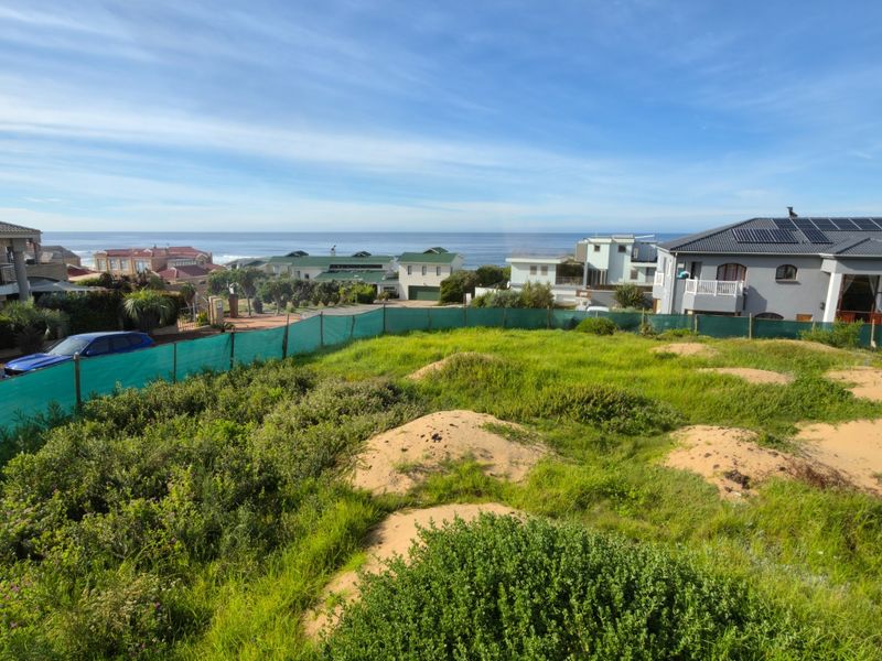 A wide-angle view of a vacant residential plot featuring natural coastal vegetation and sand dunes, overlooking a developed neighborhood and the ocean.