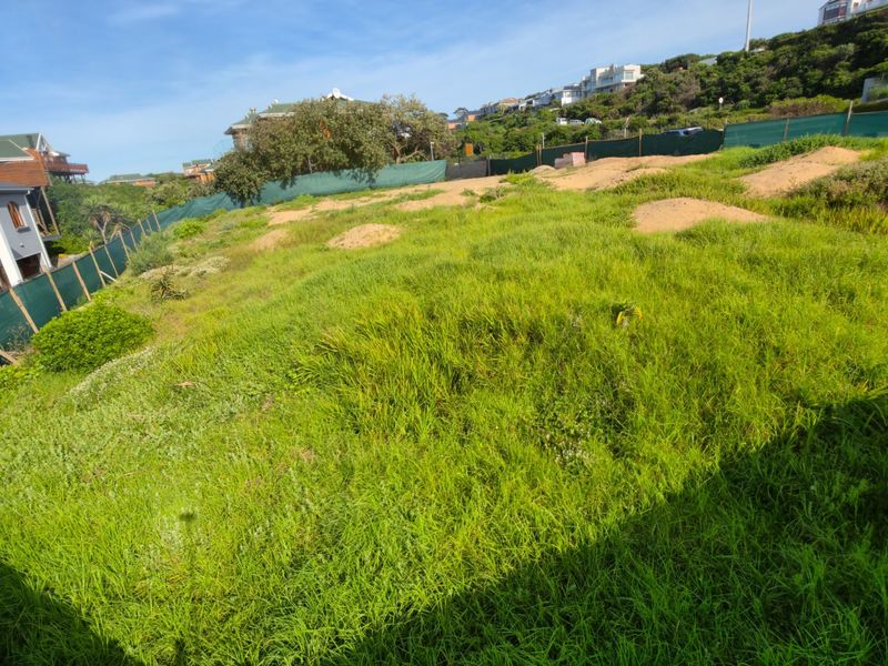 A vacant residential plot featuring sloped terrain and perimeter fencing, situated in a developed neighborhood.