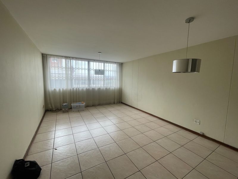 A spacious, empty living area featuring neutral tiled flooring, large windows with security bars, and modern pendant lighting.