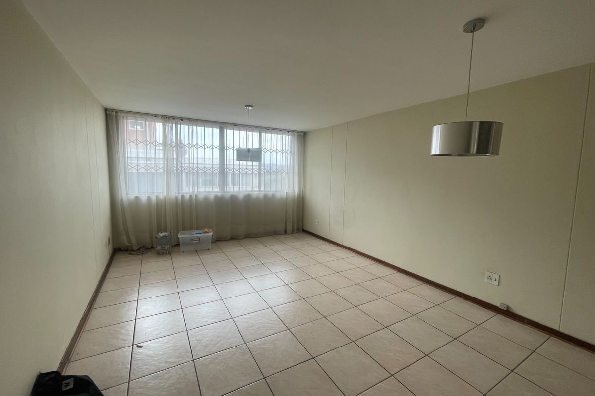 A spacious, empty living area featuring neutral tiled flooring, large windows with security bars, and modern pendant lighting.