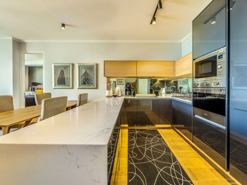 Modern open-plan kitchen featuring a stone breakfast bar, high-gloss cabinetry, integrated appliances, and sleek track lighting, seamlessly flowing into the dining area.