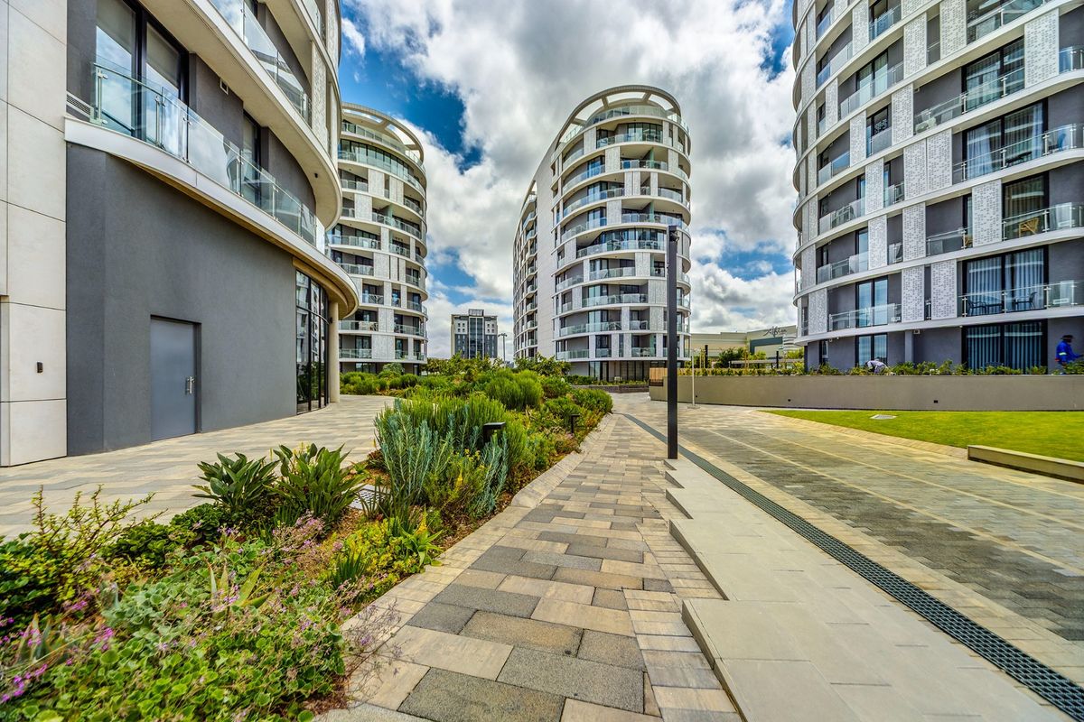 A well-maintained outdoor courtyard area between modern apartment buildings, featuring paved walkways, landscaped garden beds, and contemporary architectural design.