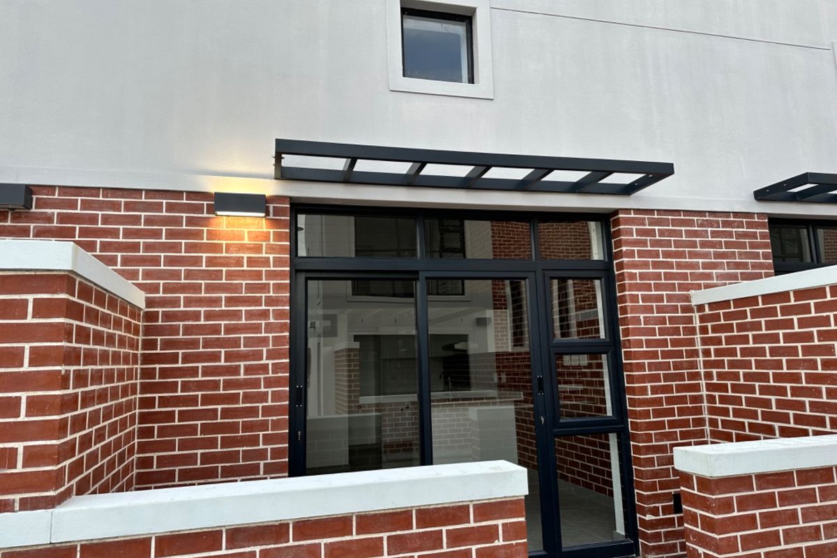 Rear exterior view of the townhouse featuring exposed brickwork, black aluminium sliding doors, a modern metal awning, and a private brick-walled courtyard area.