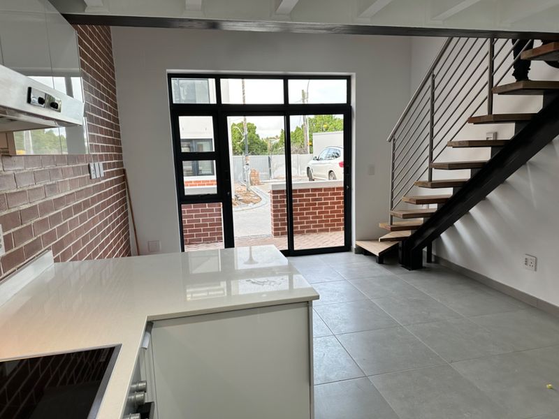 Modern open-plan living area featuring an exposed brick wall, sleek kitchen cabinetry with stone countertops, and a contemporary staircase leading to the upper level.