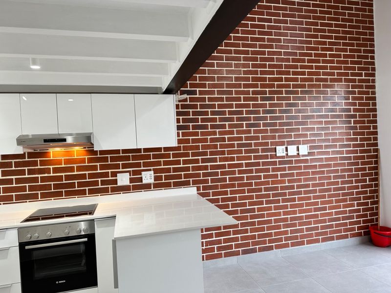 Modern kitchen featuring white cabinetry, stone countertops, integrated oven and hob, and a striking exposed brick feature wall with exposed ceiling beams.