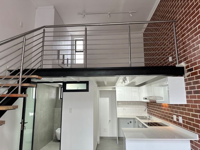 Modern open-plan living area featuring a mezzanine level with stainless steel balustrades, exposed brick accents, and a sleek, contemporary kitchen with integrated appliances.