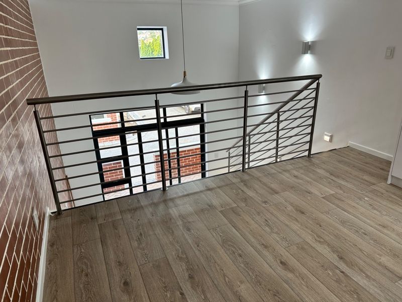 A modern landing area featuring a sleek stainless steel balustrade, wood-look laminate flooring, and an exposed brick feature wall with contemporary lighting.