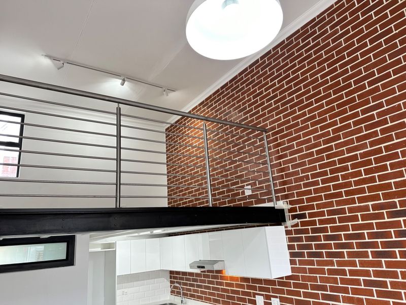 Modern open-plan living area featuring a mezzanine level with a stainless steel balustrade, exposed brick wall, and a contemporary kitchen with white cabinetry.