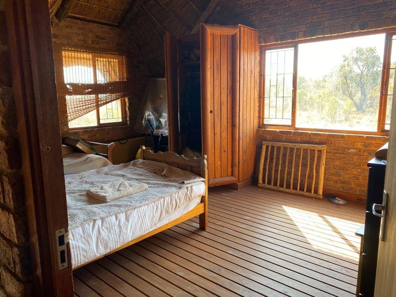 Rustic bedroom featuring exposed brick walls, a traditional thatched roof, and timber slat flooring with large windows overlooking the surrounding landscape.