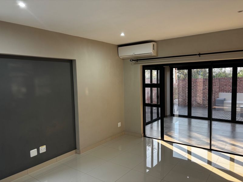 A bright, empty living room featuring tiled floors, recessed lighting, a wall-mounted air conditioner, and large black aluminium sliding doors leading to the patio.